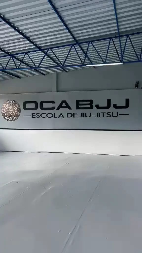 Tatame branco e parede com o letreiro OCA BJJ, Escola de Jiu-Jitsu, sob a estrutura azul do telhado